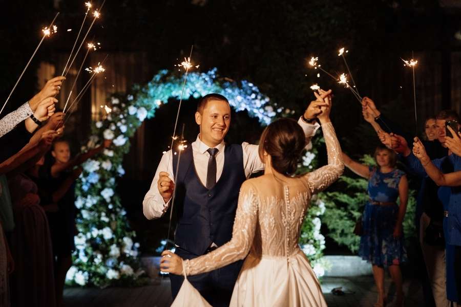 Un couple de jeunes mariés danse entouré de leurs invités tenant des cierges magiques lors d'une soirée de mariage en plein air.