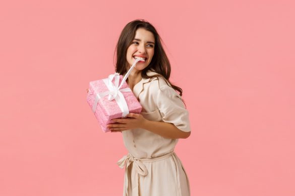 Une jeune femme souriante tient un coffret cadeau bijou femme en forme de boîte rose au ruban blanc.