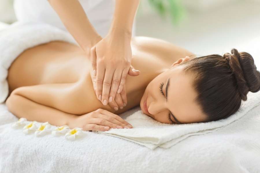 Une femme détendue, les yeux fermés, allongée sur une table de massage et recevant un massage du dos pour nourrir sa peau avec de l'huile par les mains d'un praticien.