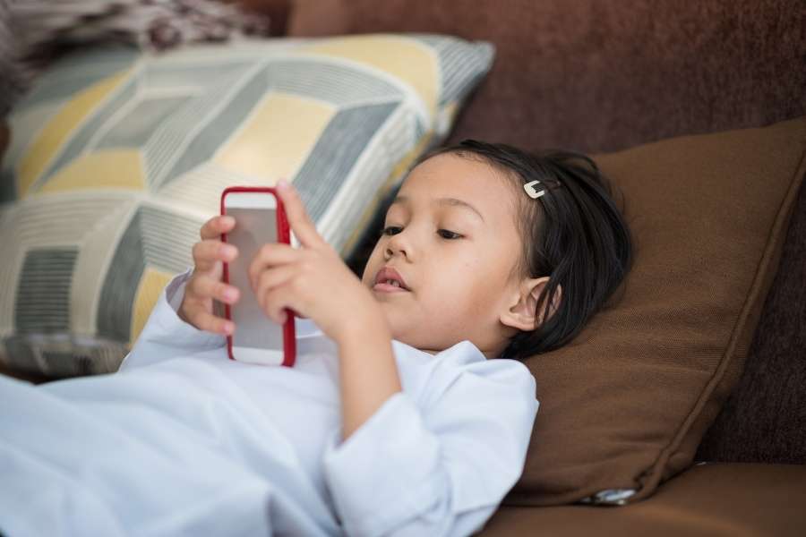 Jeune enfant allongé sur un canapé absorbé par l'écran d'un smartphone rouge.